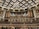 An ornate concert hall features a large pipe organ with decorative woodwork and portraits of composers above. The ceiling displays an intricate pattern with a grand chandelier at its center. Statues of figures in classical poses are placed on the sides. The lower level shows musicians and musical stands, indicating preparations for a performance.