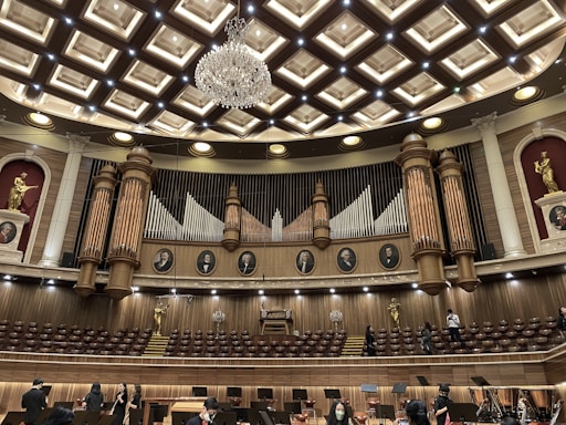 An ornate concert hall features a large pipe organ with decorative woodwork and portraits of composers above. The ceiling displays an intricate pattern with a grand chandelier at its center. Statues of figures in classical poses are placed on the sides. The lower level shows musicians and musical stands, indicating preparations for a performance.