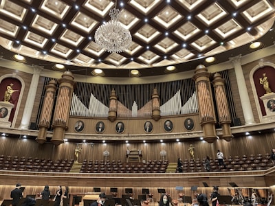 An ornate concert hall features a large pipe organ with decorative woodwork and portraits of composers above. The ceiling displays an intricate pattern with a grand chandelier at its center. Statues of figures in classical poses are placed on the sides. The lower level shows musicians and musical stands, indicating preparations for a performance.