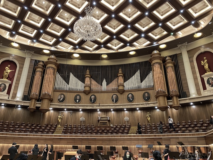 An ornate concert hall features a large pipe organ with decorative woodwork and portraits of composers above. The ceiling displays an intricate pattern with a grand chandelier at its center. Statues of figures in classical poses are placed on the sides. The lower level shows musicians and musical stands, indicating preparations for a performance.