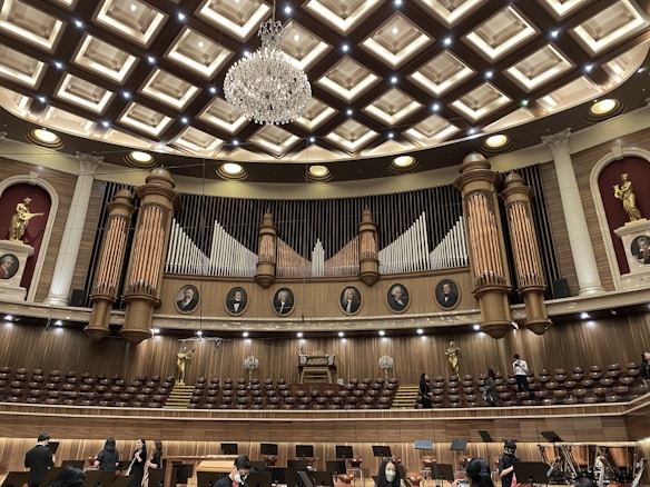An ornate concert hall features a large pipe organ with decorative woodwork and portraits of composers above. The ceiling displays an intricate pattern with a grand chandelier at its center. Statues of figures in classical poses are placed on the sides. The lower level shows musicians and musical stands, indicating preparations for a performance.