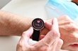 A person wearing a smartwatch on their wrist. The watch screen displays a red heart and a waveform, suggesting a health or heart rate monitoring feature. The person's hand is adjusting the watch. In the background, a blue surgical face mask is partially visible.