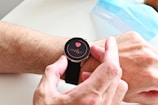 A person wearing a smartwatch on their wrist. The watch screen displays a red heart and a waveform, suggesting a health or heart rate monitoring feature. The person's hand is adjusting the watch. In the background, a blue surgical face mask is partially visible.