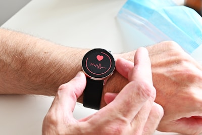 A person wearing a smartwatch on their wrist. The watch screen displays a red heart and a waveform, suggesting a health or heart rate monitoring feature. The person's hand is adjusting the watch. In the background, a blue surgical face mask is partially visible.