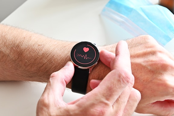 A person wearing a smartwatch on their wrist. The watch screen displays a red heart and a waveform, suggesting a health or heart rate monitoring feature. The person's hand is adjusting the watch. In the background, a blue surgical face mask is partially visible.