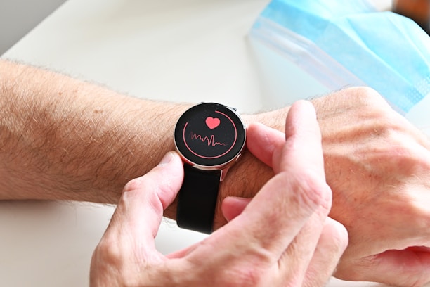 A person wearing a smartwatch on their wrist. The watch screen displays a red heart and a waveform, suggesting a health or heart rate monitoring feature. The person's hand is adjusting the watch. In the background, a blue surgical face mask is partially visible.