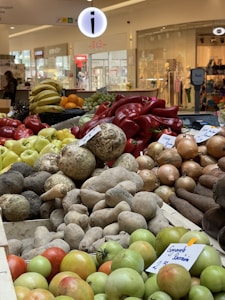 A colorful and diverse display of fruits and vegetables is arranged in a vibrant market setting. Items include different types of peppers, bananas, grapes, and root vegetables like yams and onions. The background shows a shopping mall interior with various stores and mannequins.