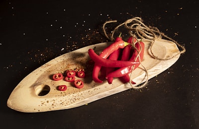 A wooden board with several whole red chili peppers and sliced chili pieces is placed on a dark surface. Twine is positioned haphazardly around the peppers, adding a rustic touch. Scattered spices and seeds are visible, enhancing the culinary theme.