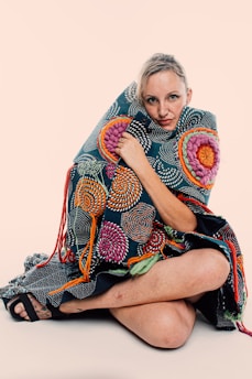 A person is seated with legs crossed, wrapped in a colorful, intricately designed textile. The fabric features swirling patterns in vibrant colors such as pink, orange, and green against a dark background. The person's expression is calm and introspective.