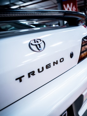 Close-up view of the back of a white car, featuring the Toyota emblem and the model name 'Trueno' in bold letters. The image is taken in what appears to be an indoor parking area, with part of a red wall with 'W6' visible in the background.
