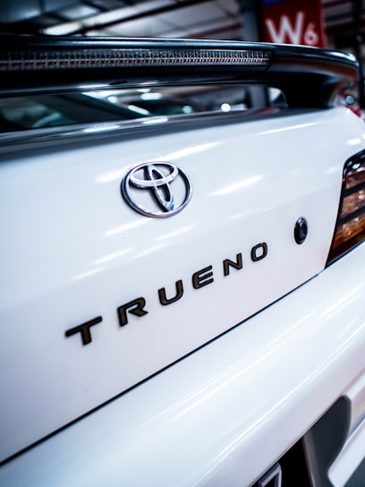 Close-up view of the back of a white car, featuring the Toyota emblem and the model name 'Trueno' in bold letters. The image is taken in what appears to be an indoor parking area, with part of a red wall with 'W6' visible in the background.