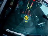 The interior of a car is visible through the windshield, prominently featuring a steering wheel with a distinctive yellow center and brand logo. Racing harnesses with red straps are partially visible in the background.