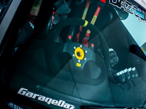 The interior of a car is visible through the windshield, prominently featuring a steering wheel with a distinctive yellow center and brand logo. Racing harnesses with red straps are partially visible in the background.