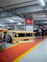 A vintage car with a customized body kit and yellow headlights is parked in a well-lit underground parking garage. The vehicle is positioned in front of a red and yellow painted floor, next to other classic cars. Structural beams and bright ceiling lights are visible above.