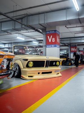 A vintage car with a customized body kit and yellow headlights is parked in a well-lit underground parking garage. The vehicle is positioned in front of a red and yellow painted floor, next to other classic cars. Structural beams and bright ceiling lights are visible above.