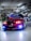 A customized red BMW car is parked in an indoor garage, featuring vibrant headlight modifications. The hood is adorned with graphics, and other cars with elaborate designs are visible in the background. The garage has numerous overhead lights and visible piping.