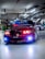 A customized red BMW car is parked in an indoor garage, featuring vibrant headlight modifications. The hood is adorned with graphics, and other cars with elaborate designs are visible in the background. The garage has numerous overhead lights and visible piping.