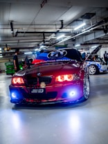 A customized red BMW car is parked in an indoor garage, featuring vibrant headlight modifications. The hood is adorned with graphics, and other cars with elaborate designs are visible in the background. The garage has numerous overhead lights and visible piping.