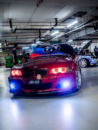 A customized red BMW car is parked in an indoor garage, featuring vibrant headlight modifications. The hood is adorned with graphics, and other cars with elaborate designs are visible in the background. The garage has numerous overhead lights and visible piping.