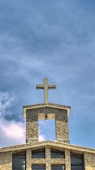 A church building with a mosaic-patterned facade and a prominent cross on the roof. The sky is partly cloudy with a blue hue, and large windows are visible on the front of the structure.