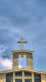 A church building with a mosaic-patterned facade and a prominent cross on the roof. The sky is partly cloudy with a blue hue, and large windows are visible on the front of the structure.