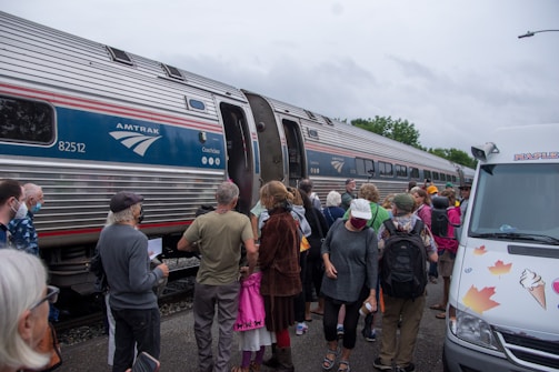 A group of people stand near an Amtrak train, with some individuals preparing to board. The scene includes a variety of people wearing casual clothing, some holding bags or papers. Nearby, a van decorated with maple leaf and ice cream cone illustrations is parked.