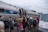 A group of people stand near an Amtrak train, with some individuals preparing to board. The scene includes a variety of people wearing casual clothing, some holding bags or papers. Nearby, a van decorated with maple leaf and ice cream cone illustrations is parked.
