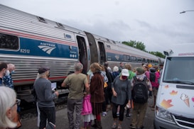 A group of people stand near an Amtrak train, with some individuals preparing to board. The scene includes a variety of people wearing casual clothing, some holding bags or papers. Nearby, a van decorated with maple leaf and ice cream cone illustrations is parked.