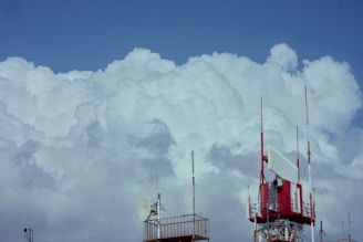 A large, white antenna tower with several transmitters and dishes stands against a backdrop of massive, billowing white clouds. The sky is a clear, vibrant blue, highlighting the contrast between the tower's red and white structure and the soft clouds.