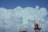 A large, white antenna tower with several transmitters and dishes stands against a backdrop of massive, billowing white clouds. The sky is a clear, vibrant blue, highlighting the contrast between the tower's red and white structure and the soft clouds.