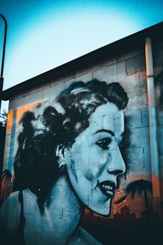 A large mural painted on a concrete wall features a detailed and artistic depiction of a woman's side profile. The artwork uses shades of blue and black, with touches of white for highlights. The woman's expression is peaceful and slightly smiling. The background shows palm trees and a silhouette of a cityscape.