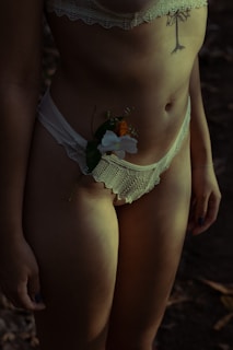 A person wearing lace lingerie with a tattoo on the torso. A delicate flower is tucked into the waistband. The lighting is dim, creating shadows and a moody atmosphere.