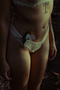 A person wearing lace lingerie with a tattoo on the torso. A delicate flower is tucked into the waistband. The lighting is dim, creating shadows and a moody atmosphere.