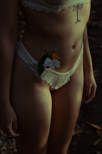 A person wearing lace lingerie with a tattoo on the torso. A delicate flower is tucked into the waistband. The lighting is dim, creating shadows and a moody atmosphere.