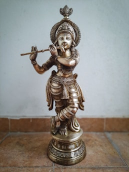 A detailed and intricately designed bronze statue depicting a figure playing a flute. The statue features elaborate ornamentation and stands on a decorated pedestal. The figure is garbed in traditional attire with meticulously crafted jewelry. The posture suggests a sense of grace and movement.