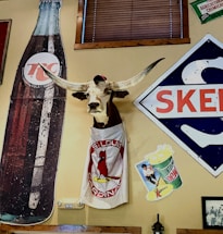 A decor featuring a mounted longhorn head wearing a cap and a T-shirt with the St. Louis Cardinals logo. Nearby are vintage advertisements including a large 7up bottle, a Mountain Dew poster, and a Skelly sign. The setting has a nostalgic, rustic ambiance with wooden elements.