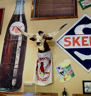 A decor featuring a mounted longhorn head wearing a cap and a T-shirt with the St. Louis Cardinals logo. Nearby are vintage advertisements including a large 7up bottle, a Mountain Dew poster, and a Skelly sign. The setting has a nostalgic, rustic ambiance with wooden elements.