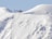 A red helicopter is hovering above a snow-covered mountainous landscape. The snow appears to be pristine and untouched, creating a serene and remote atmosphere. The silhouette of the helicopter stands out vividly against the soft, white background of the snow and the gentle slopes of the mountains.