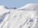 A red helicopter is hovering above a snow-covered mountainous landscape. The snow appears to be pristine and untouched, creating a serene and remote atmosphere. The silhouette of the helicopter stands out vividly against the soft, white background of the snow and the gentle slopes of the mountains.
