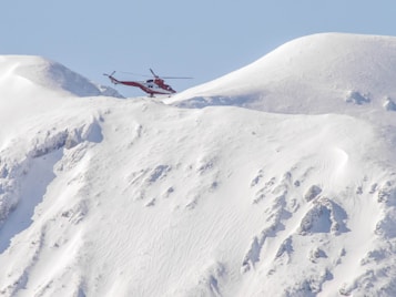 A red helicopter is hovering above a snow-covered mountainous landscape. The snow appears to be pristine and untouched, creating a serene and remote atmosphere. The silhouette of the helicopter stands out vividly against the soft, white background of the snow and the gentle slopes of the mountains.