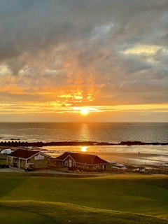 A serene seaside view at sunset with golden hues casting a warm glow over the ocean. The sun is partially hidden by clouds, creating a dramatic sky. In the foreground, there are small beachside buildings and a stretch of grassy land.