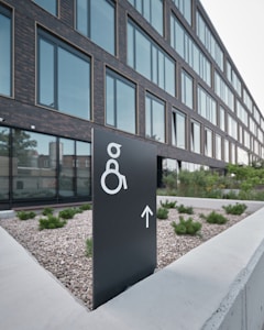 A modern building with large glass windows is seen in the background. In the foreground, there is a black sign with a white symbol for wheelchair accessibility and an arrow pointing forward. The sign is placed on a concrete surface surrounded by small shrubs and gravel.