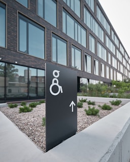 A modern building with large glass windows is seen in the background. In the foreground, there is a black sign with a white symbol for wheelchair accessibility and an arrow pointing forward. The sign is placed on a concrete surface surrounded by small shrubs and gravel.
