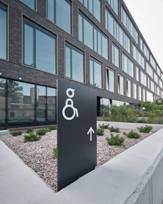 A modern building with large glass windows is seen in the background. In the foreground, there is a black sign with a white symbol for wheelchair accessibility and an arrow pointing forward. The sign is placed on a concrete surface surrounded by small shrubs and gravel.