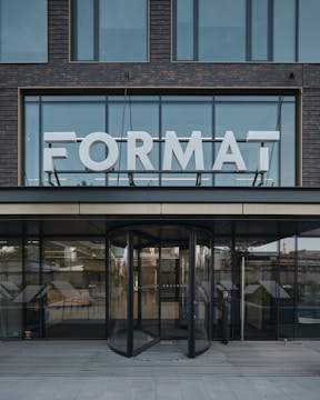 A modern building entrance featuring large glass windows and a revolving door. The architecture includes a prominent sign displaying the word 'FORMAT' in bold, white letters. The exterior is built with dark brick and metal elements, creating a sleek, contemporary look.