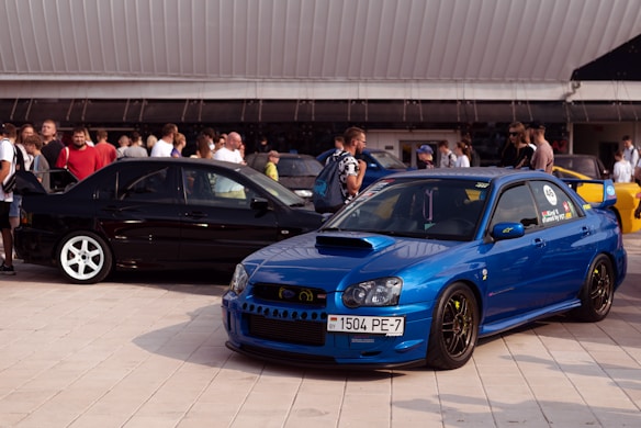 A car meet event with two prominently featured cars in the foreground. The car on the right is a blue sports car with a spoiler and racing stickers on its windows. The car on the left is a black vehicle with white wheels. A crowd of people is gathered around, some appearing to engage in conversations, and the setting is an outdoor area with a modern architectural backdrop.