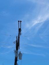 A wooden utility pole stands tall against a clear blue sky. The pole is equipped with several crossarms and multiple electrical transformers. Numerous power lines extend from the top and middle parts of the pole, stretching across the frame. The upper portions of the pole have two black insulators or equipment pieces.