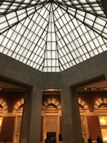 An interior view of a building with a high, geometric glass ceiling in a grid pattern. Below, there are tall arched doorways with a mix of natural and artificial lighting. The architecture includes a combination of modern and classic design elements.