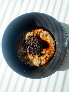 A black bowl contains a serving of oatmeal topped with a dark sauce or condiment in the center, which appears glossy due to the lighting. The bowl is placed on a white surface with diagonal shadows cast across it.