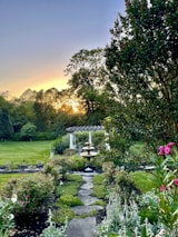 A tranquil garden scene with a stone pathway leading to a multi-tiered fountain. Lush greenery and colorful flowers surround the area, creating a serene and picturesque environment. A white pergola is visible in the background, positioned under large, leafy trees as the sun sets, casting a warm glow.